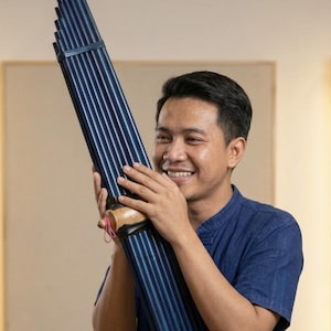 Puede incluir: Un instrumento musical de boca tradicional que presenta múltiples tubos de bambú largos de color azul oscuro dispuestos en una fila vertical, conectados por una cámara de aire central de madera con un pequeño lazo de hilo rosa.