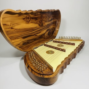 Pode incluir: Um saltério de madeira com um estojo de madeira a condizer. O saltério tem uma caixa de ressonância amarelo claro com cordas e uma moldura de madeira castanha escura. O estojo está aberto, revelando o instrumento. Instrumento musical.