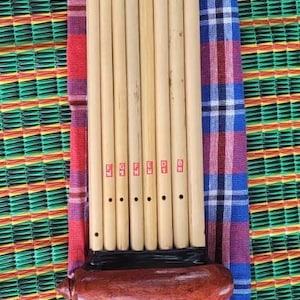 Puede incluir: Flauta de pan de bambú hecha a mano, un instrumento de viento tradicional. El instrumento tiene múltiples tubos de bambú de diferentes longitudes, unidos. Descansa sobre una tela a cuadros colorida y una estera tejida.