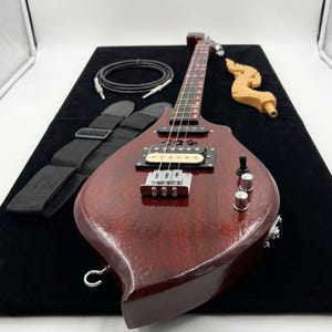Könnte beinhalten: Eine dunkelrote E-Gitarre mit einem einzigartigen, tropfenförmigen Korpus. Die Gitarre verfügt über einen schwarzen Gurt, ein schwarzes Kabel und ein dekoratives Holzelement. Das Griffbrett hat rote Akzente. Die Gitarre liegt auf einer schwarzen Oberfläche.