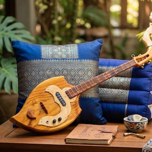 May include: A traditional wooden stringed instrument with a decorative headstock. It is placed on a wooden table, with two blue cushions with gold patterned fabric behind it. A small bowl, notebook, and fabric are also on the table.