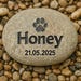 May include: A light grey, oval stone engraved with the name "Honey" in black, bold lettering. Above the name is a paw print, and below is the date "21.05.2025". The stone is surrounded by small, smooth pebbles.