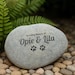 May include: A grey memorial stone, inscribed with "In Loving Memory of Opie & Lila" and two paw prints. The oval-shaped stone sits on a bed of small, rounded stones, with green foliage in the background.