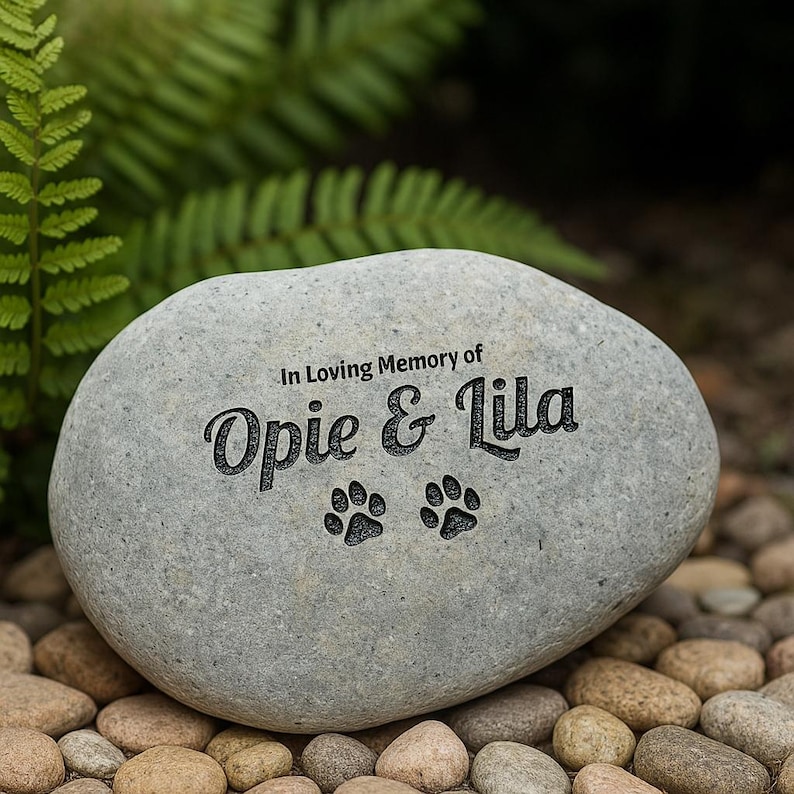 May include: A grey memorial stone, inscribed with "In Loving Memory of Opie & Lila" and two paw prints. The oval-shaped stone sits on a bed of small, rounded stones, with green foliage in the background.