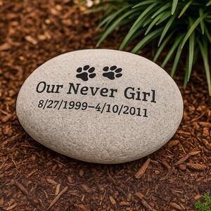 May include: A grey, oval memorial stone with black paw prints and the inscription "Our Never Girl" and dates 8/27/1999-4/10/2011. The stone is set in a bed of brown mulch with green plants in the background.