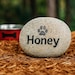 May include: A light grey stone engraved with the name "Honey" and a paw print. The stone sits on brown mulch, with a red dog bowl in the background. A pet-themed garden ornament.