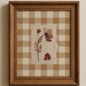 Könnte beinhalten: Gerahmtes Kunstwerk mit einem Arrangement aus gepressten Blumen. Das Kunstwerk ist vor einem karierten Hintergrund in Beige und Weiß platziert. Der Rahmen ist aus Holz. Die gepressten Blumen sind in Lila- und Rottönen gehalten.