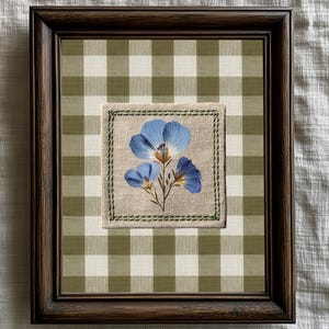 Op de afbeelding: Een ingelijst kunstwerk met een botanische print van blauwe bloemen. Het kunstwerk is geplaatst tegen een groen-witte geruite achtergrond. De lijst is donkerbruin.