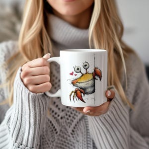 May include: White ceramic mug featuring a cartoon crab design. The crab has a yellow body, red claws, and large, googly eyes. Two small red hearts are next to the crab. The mug has a white handle.