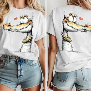 May include: White t-shirt featuring a whimsical illustration of a smiling alligator with a heart. The alligator has yellow and orange accents. The design is on the front and back of the shirt. The shirt is a casual, everyday wear item.