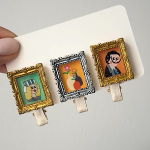 May include: Three miniature art-themed hair clips. Each clip features a different framed image: a woman in a hat, a vase of flowers, and a man with a mustache. The frames are gold and silver, with cream-coloured ribbon clips.