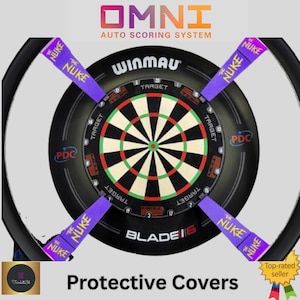 May include: A black dartboard with a green, red, and white target design. The board has the text "WINMAU" and "BLADE 6". Four purple dart protectors with "THE NUKE" text are placed around the board. The text "OMNI AUTO SCORING SYSTEM" is at the top.