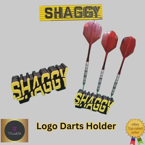 May include: A darts holder set with the word "SHAGGY" in yellow and black. The set includes a darts holder with three darts and a separate "SHAGGY" logo. The darts have red flights.