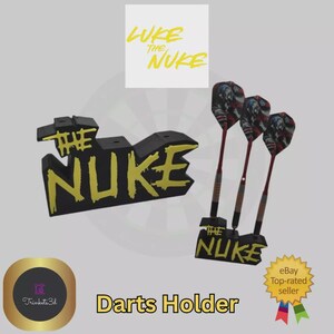 May include: Black and yellow "The Nuke" darts holder with three darts. The darts have red shafts and American flag-themed flights. The text "Luke the Nuke" is above the holder. The words "Darts Holder" are at the bottom.