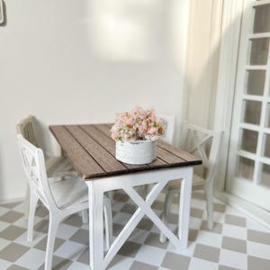 Op de afbeelding: Witte eettafel in landelijke stijl met twee witte stoelen en een witte plantenbak met roze bloemen op de tafel. De tafel heeft een houten blad en een witte basis. De vloer is een zwart-wit geblokt patroon.