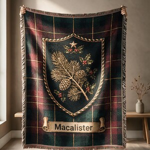 Op de afbeelding: Een geweven plaid met een donkergroen en rood ruitpatroon en een centraal schildembleem. Het schild toont een gouden dennentak, dennenappels, hulstbessen en een ster. Een scroll aan de onderkant bevat de naam Macalister.