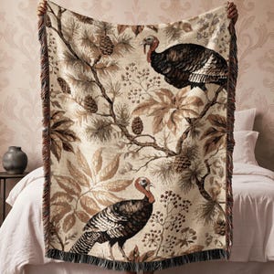 May include: A woven throw blanket featuring a detailed pattern of two turkeys, pine branches, pinecones, and foliage in shades of brown, tan, and cream. The rectangular blanket has a decorative fringed edge along the sides and bottom.
