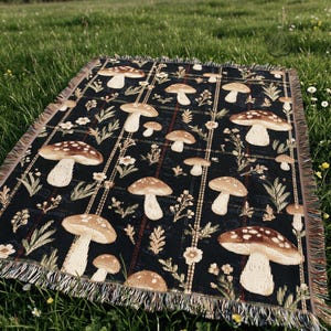 May include: A woven throw blanket featuring a repeating pattern of brown and white mushrooms, small flowers, and green foliage on a black background with a plaid grid overlay and frayed edges along all four sides of the rectangular textile.