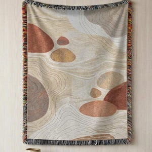 May include: A woven throw blanket featuring an abstract pattern of smooth, rounded shapes in shades of terracotta, tan, and cream. The design includes fine, wavy lines resembling sand ripples, and the edges are finished with a multicolored fringe.