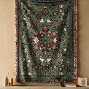 May include: A rectangular woven throw blanket featuring a dark green background with a symmetrical floral pattern in shades of deep red, cream, and sage green. The blanket has a decorative fringed edge along all four sides.