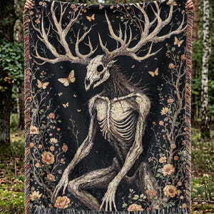 May include: Black throw blanket featuring a skeletal figure with antlers, surrounded by floral and butterfly motifs. The artwork has a dark, gothic aesthetic with a fringed edge. The color palette includes black, beige, and brown.