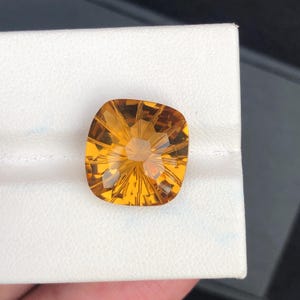 Puede incluir: Una gema facetada de color amarillo dorado con talla cojín cuadrada. La gema se encuentra sobre un fondo blanco, mostrando sus intrincados patrones internos y brillo. El color de la gema varía de ámbar claro a oscuro.