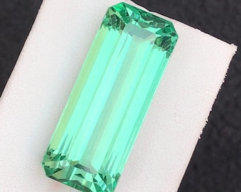 May include: A rectangular, faceted gemstone with a vibrant, light green hue. The gem is set against a white surface, showcasing its clarity and brilliance. The cut of the stone allows light to pass through, creating a sparkling effect.