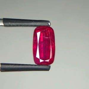Puede incluir: Un rubí de corte rectangular, sujeto por unas pinzas negras. El rubí es de color rojo intenso con facetas internas que reflejan la luz. La gema se muestra sobre un fondo gris neutro. Medidas en milímetros.