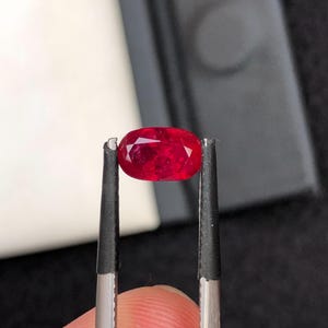 Puede incluir: Una gema roja de corte ovalado sostenida por unas pinzas de metal. La piedra presenta una superficie facetada que refleja la luz, mostrando un tono carmesí profundo y saturado en toda su estructura, con una apariencia clara y pulida.