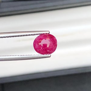 May include: A vibrant, oval-shaped ruby gemstone, held by silver tweezers. The ruby displays a rich, deep red color with internal light reflections. The gemstone is smooth and polished, showcasing its natural beauty. Ideal for jewelry making.
