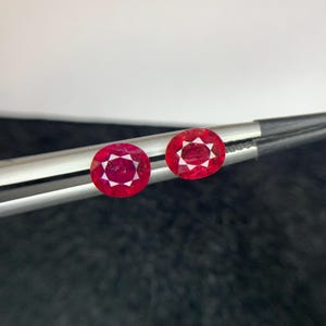 May include: Two oval-cut, vibrant red rubies held by silver-toned metal tweezers. Each gemstone features a faceted surface that reflects light, showcasing a deep crimson hue and a clear, polished appearance against a dark, textured background.