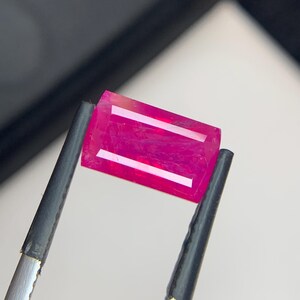 Peut inclure: Un rubis de coupe rectangulaire, maintenu par une pince. Le rubis affiche une teinte rouge-rosé profond avec deux traits internes parallèles plus clairs. La gemme est tenue sur un fond neutre, soulignant sa couleur et sa clarté.