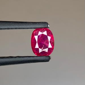 Może przedstawiać: Owalny rubin trzymany czarną pęsetą. Rubin ma głęboki czerwony odcień z białym wzorem w kształcie gwiazdy pośrodku. Kamień szlachetny jest umieszczony na neutralnym tle, podkreślając jego kolor i szlif.
