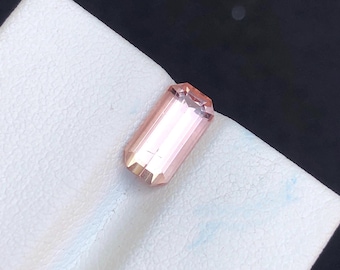 May include: A rectangular, faceted gemstone with a light peach color. The gem is set against a white surface, showcasing its polished surfaces and internal clarity. The gemstone's cut allows light to reflect, creating a subtle sparkle.