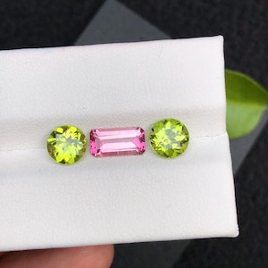 Puede incluir: Tres piedras preciosas expuestas sobre una superficie blanca. Dos piedras redondas y facetadas de peridoto verde claro flanquean una gema rectangular de turmalina rosa. Las gemas se presentan sobre un fondo neutro, resaltando su color y talla.