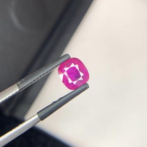 May include: A vibrant, cushion-cut ruby gemstone held by tweezers. The ruby displays a deep pinkish-red hue with a starburst pattern in the center. The gemstone is held against a white and black background.