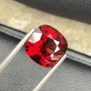 Puede incluir: Una gema roja de talla cojín sostenida por unas pinzas metálicas de tono plateado. La piedra presenta una superficie facetada que refleja la luz, creando reflejos de color carmesí profundo y escarlata brillante en su estructura.
