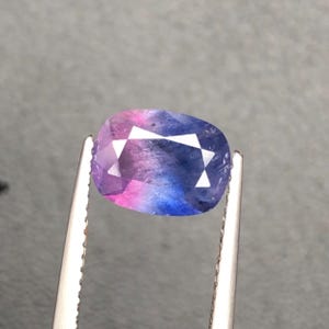 Może przedstawiać: Wielokolorowy kamień szlachetny z mieszanką różowych, fioletowych i niebieskich odcieni. Kamień szlachetny jest trzymany przez srebrną pęsetę, prezentując jego wypolerowane fasetki i unikalny gradient kolorów. Kamień ma owalny kształt.