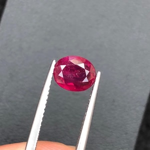 Könnte beinhalten: Ein leuchtender, oval geschliffener Edelstein in tiefem Rot mit zahlreichen dreieckigen Facetten, die das Licht reflektieren. Der Stein wird von einer silberfarbenen Metallpinzette gehalten und zeigt seine Transparenz und polierte Oberfläche.