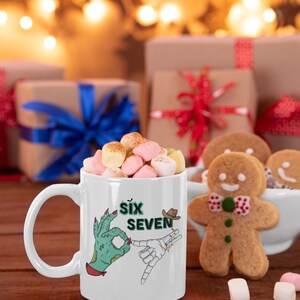 May include: White ceramic mug filled with marshmallows, featuring a graphic of two hands, one green and one skeletal, with the text "Six Seven". A gingerbread man and wrapped gifts are in the background.
