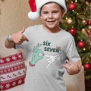 May include: A light gray t-shirt featuring a graphic with the words "SIX SEVEN" and illustrations of hands making hand gestures. The shirt is worn by a person wearing a Santa hat.