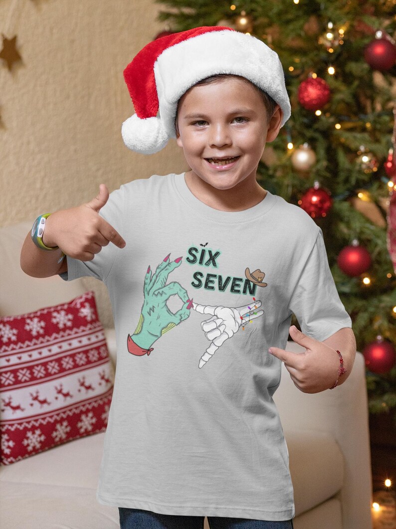 May include: A light gray t-shirt featuring a graphic with the words "SIX SEVEN" and illustrations of hands making hand gestures. The shirt is worn by a person wearing a Santa hat.