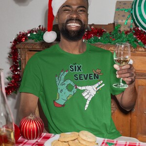 May include: Green t-shirt with the text "Six Seven" and a graphic of two hands making hand gestures. The person in the image is wearing a Santa hat and holding a glass of wine. A plate of cookies sits on a red and white checkered tablecloth.