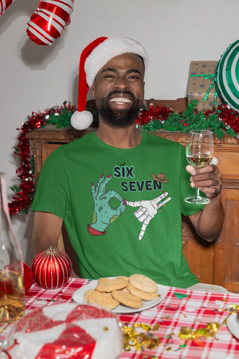 May include: Green t-shirt with the text "Six Seven" and a graphic of two hands making hand gestures. The person in the image is wearing a Santa hat and holding a glass of wine. A plate of cookies sits on a red and white checkered tablecloth.