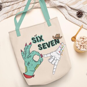 May include: Beige tote bag with teal handles featuring a Halloween-themed design. The design includes the text "SIX SEVEN", a cowboy hat, and two hands, one green and zombie-like, the other a skeleton hand.
