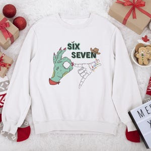 May include: White sweatshirt with a graphic featuring a green hand making an 'okay' sign and a skeleton hand. The text 'SIX SEVEN' is above the hands, with a cowboy hat. Christmas decorations surround the sweatshirt.