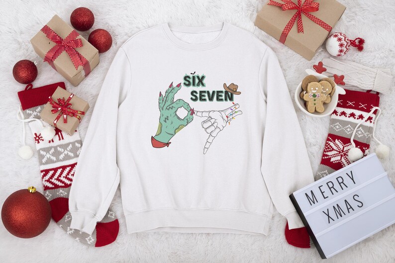 May include: White sweatshirt with a graphic featuring a green hand making an 'okay' sign and a skeleton hand. The text 'SIX SEVEN' is above the hands, with a cowboy hat. Christmas decorations surround the sweatshirt.