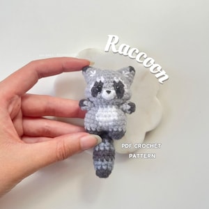 Puede incluir: Un juguete de amigurumi de mapache tejido a crochet con un cuerpo gris y blanco y una cola negra y gris. El juguete está sentado sobre una superficie blanca con el texto "Raccoon PDF CROCHET PATTERN" debajo.
