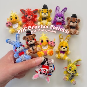 May include: A selection of vibrant, hand-crocheted plush toys, featuring yellow, red, brown, purple, and blue characters. The text "PDF Crochet Pattern fnaf 1-3 mini plushies" is displayed. The toys are arranged on a light-coloured surface.