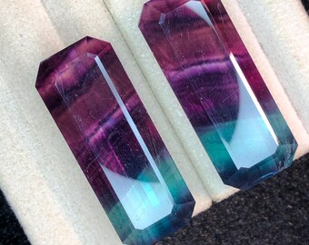 May include: Two rectangular fluorite crystals with faceted edges. The crystals display a gradient of colours, including purple, teal, and clear. The crystals are displayed on a white and grey surface.